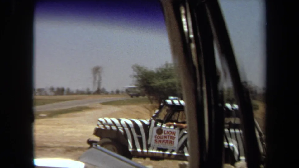 A partial 1971 home-movie view shows the striped safari Jeep near a paved road, obscured by the filming vehicle's window pillar.