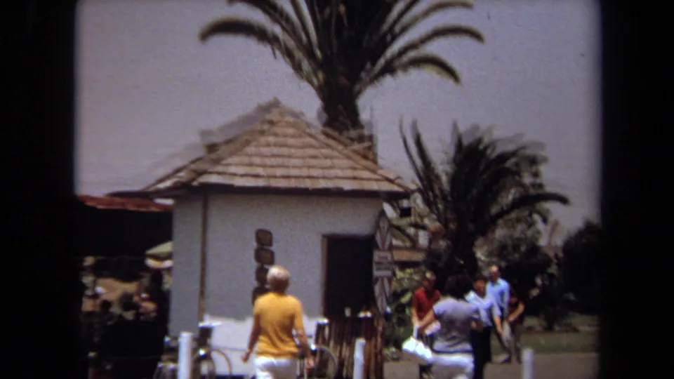 1971 Laguna Hills home-movie frame showing visitors near palm trees and a small white thatched-roof building.