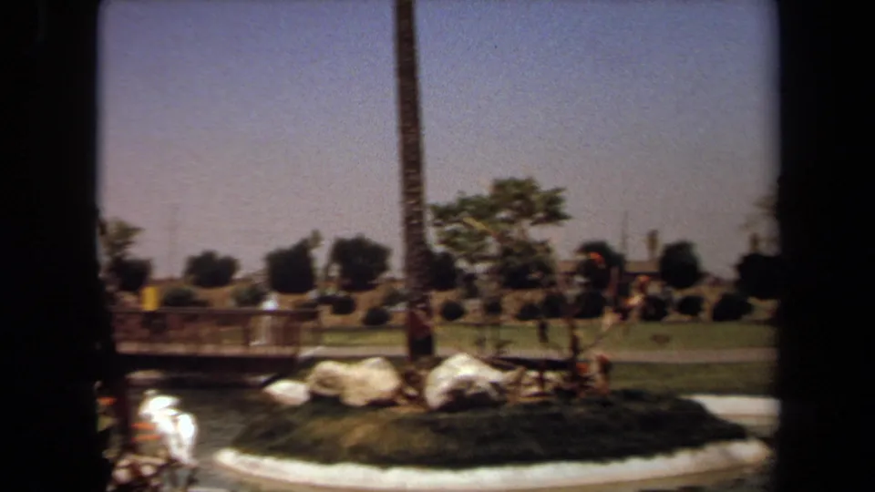 1971 home-movie frame of a Laguna Hills park pond with reflected sunlight, bridge railings, rocks, palms, grass, and distant people.