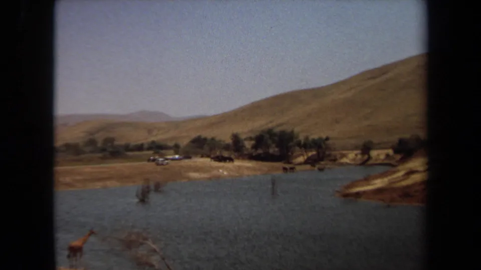 1971 Laguna Hills 8mm wide frame with water, arid hills, sparse trees, distant vehicles, and a small giraffe near the foreground.
