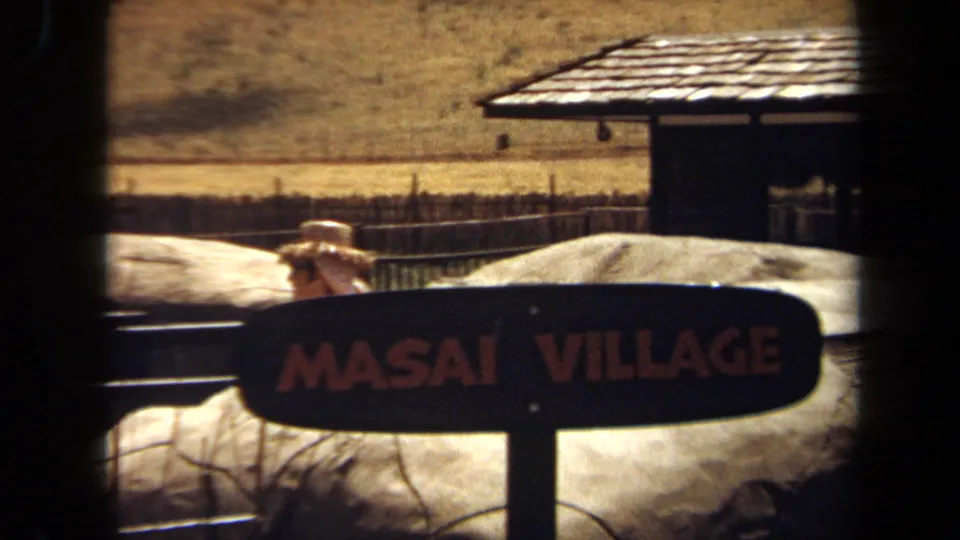 A readable Masai Village sign appears in the safari park area, with fencing, a shelter roof, and a distant visitor visible.