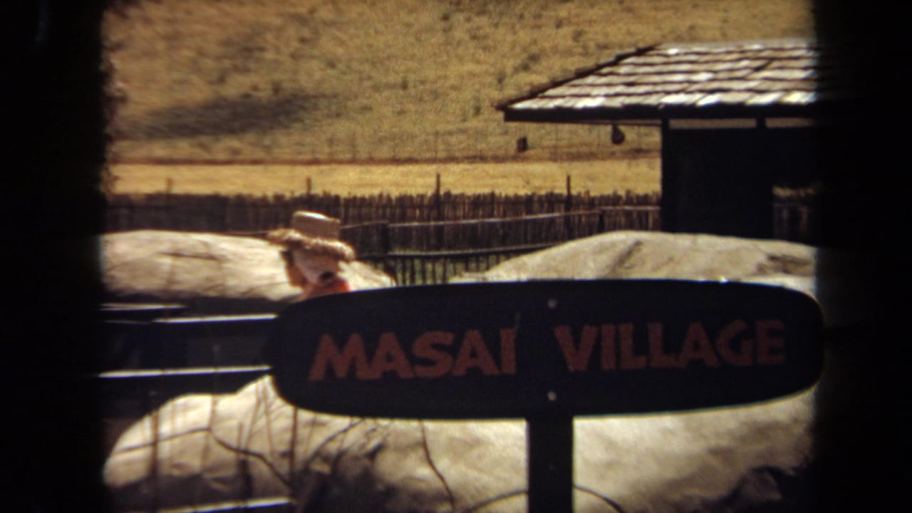 1971 Laguna Hills 8mm frame showing a woman in a straw hat near a MASAI VILLAGE exhibit sign, fencing, rocks, and a thatched structure.