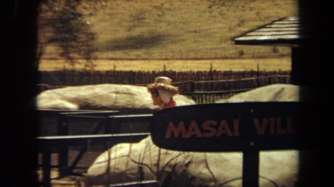Woman in a straw hat partly hidden behind the MASAI VILLAGE sign and railing.