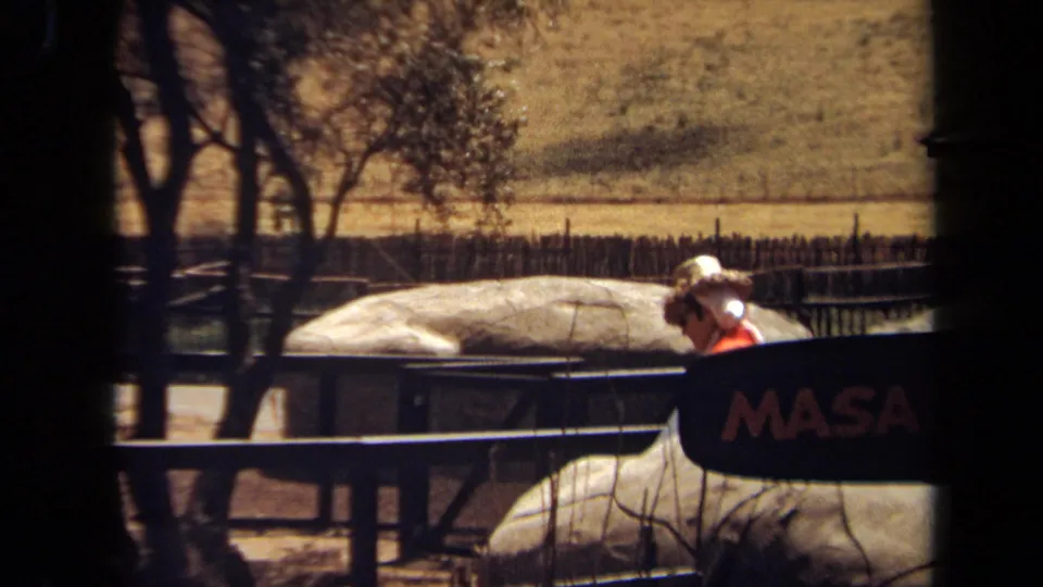 1971 8mm frame from Laguna Hills showing a visitor in a straw hat near trees, rails, pale rocks, and the edge of the MASAI VILLAGE sign.