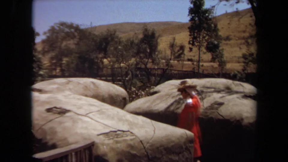 A woman in orange moves past a large rock enclosure in the 1971 Laguna Hills zoo footage, with trees and dry hills in the background.