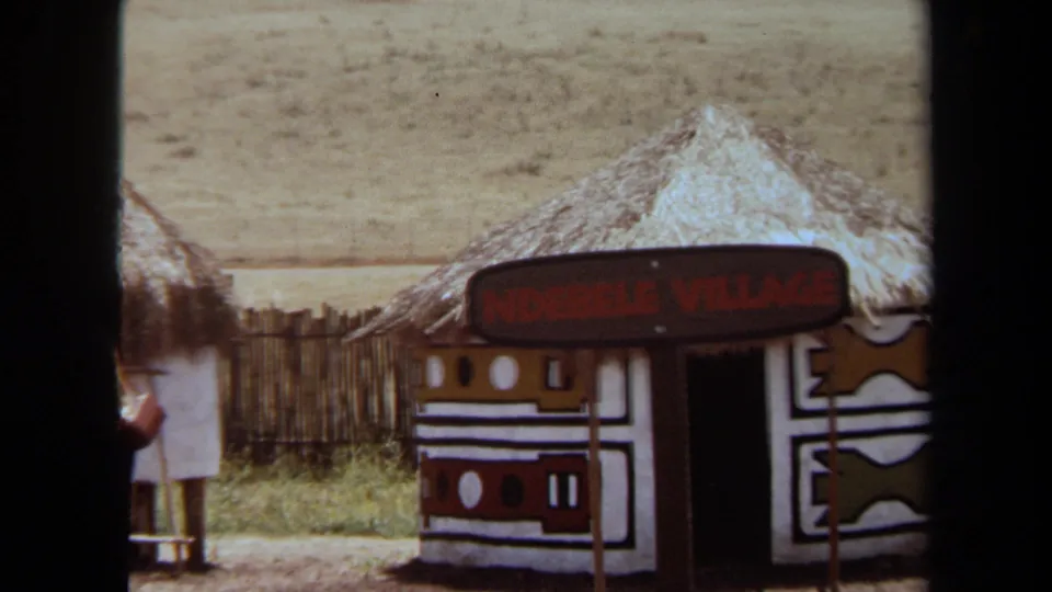A 1971 Laguna Hills frame shows the Ndebele Village exhibit hut beside fencing, with a visitor partly visible near the left edge.