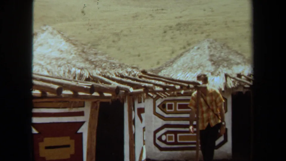 1971 Laguna Hills frame of a man beside a recreated village structure with thatched roofing, painted wall panels, and open dry ground.