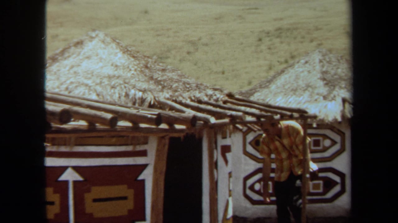 Man in a patterned shirt approaching a thatched-roof building with geometric wall designs.