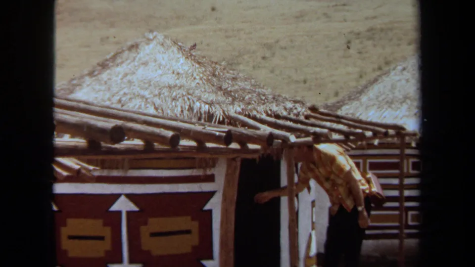 An adult man enters a shadowed doorway in a thatched-roof structure, with patterned wall panels visible in the California home-movie frame.