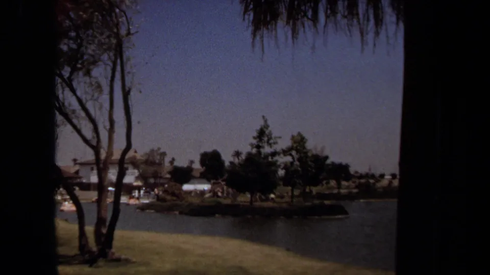 Laguna Hills home-movie footage cuts to a landscaped pond or water feature with trees, grass, and low buildings beyond it.