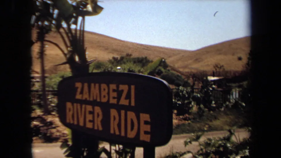 1971 8mm frame showing a Zambezi River Ride sign at a California attraction, with dry hills and foliage behind it.