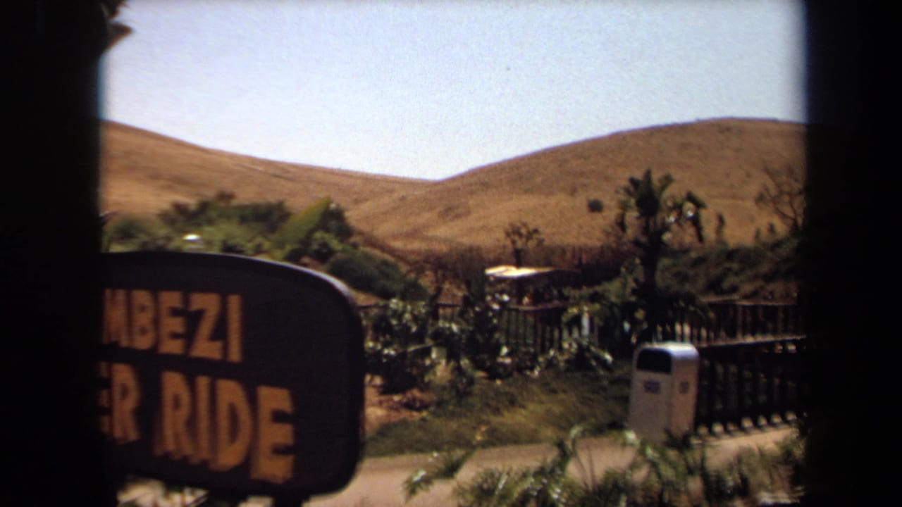 Partially obscured Zambezi River Ride sign beside fenced landscaping and hills.