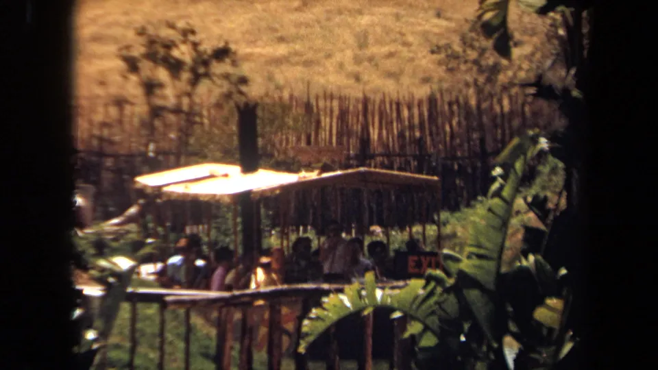 1971 8mm view of guests seated in a covered Zambezi River Ride boat beside tropical foliage and fencing.