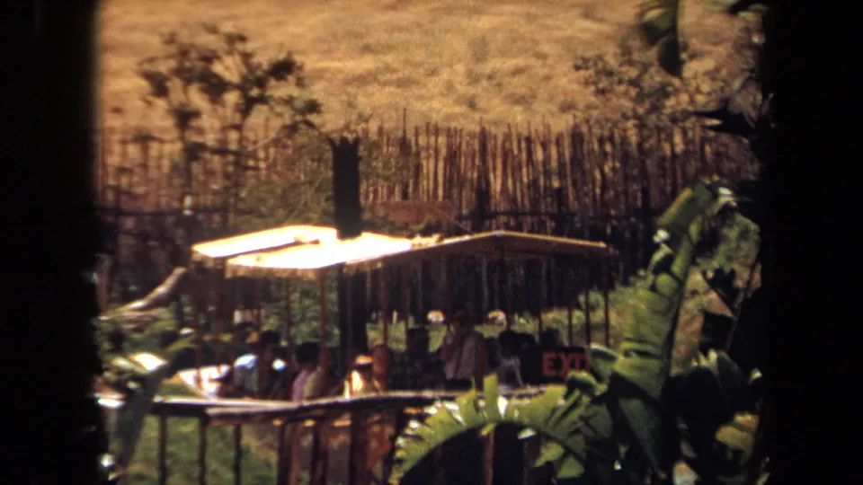 1971 Laguna Hills home-movie frame showing a canopy-covered boat with passengers moving past fencing and dense landscaped plants.