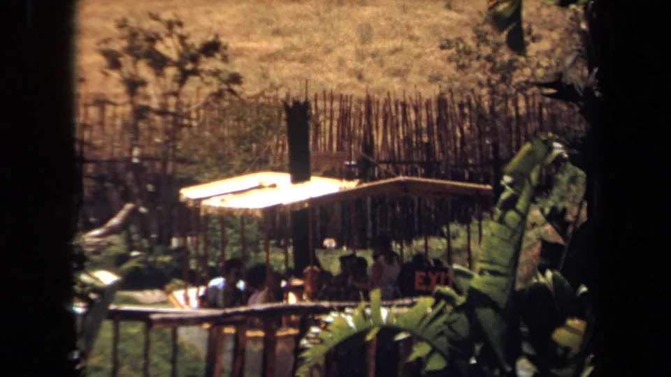 1971 Laguna Hills 8mm frame with passengers seated on a covered boat beside rustic fencing, greenery, and a dry sunlit hillside.