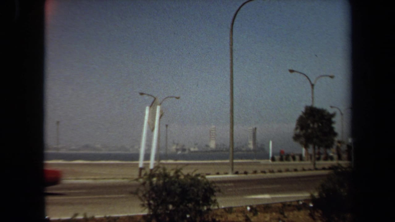 Coastal road with a red car, streetlights, shrubs, water, and distant buildings under hazy sky.