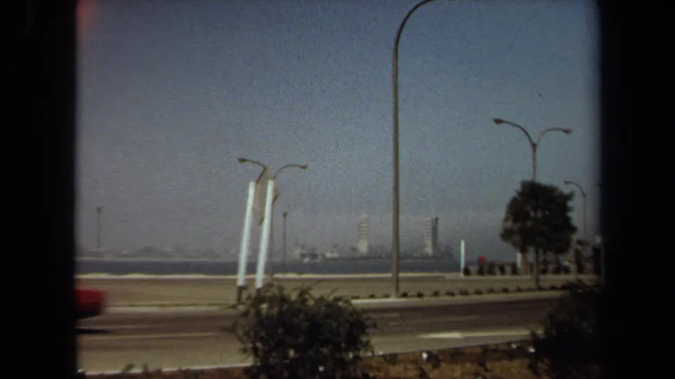 1971 8mm view from Laguna Hills metadata showing a coastal road, red car, streetlights, water, and a distant skyline.