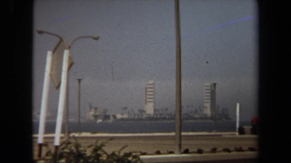 1971 8mm frame showing roadside lamps in the foreground and a hazy waterfront skyline across the water.