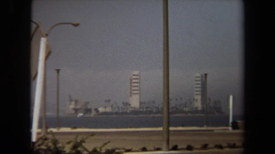 Closer 1971 8mm view toward distant high-rises and palms across the water, partly framed by roadside poles.