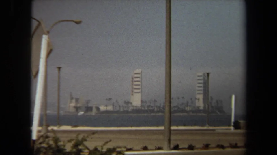 1971 8mm frame holds on the distant waterfront skyline, with palms and towers seen beyond roadside poles and shrubs.