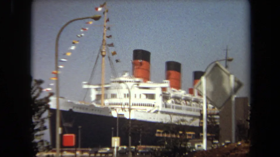 A docked ocean liner in 1971 California footage appears behind a curved streetlight, signal flags, a road sign, parked cars, and harbor-side trees.