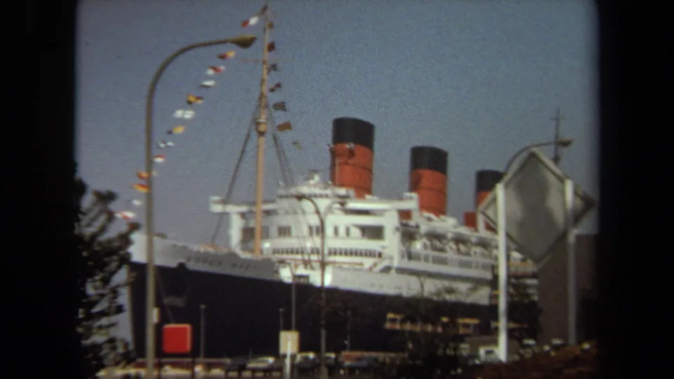 The ship's upper decks and red-and-black funnels fill the frame, seen past a streetlight, signpost, trees, and parked cars in home-movie footage.