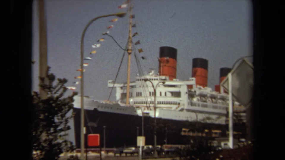 A street-level 8mm view shows the docked ocean liner with flags and funnels, partially obscured by roadside signs, lights, cars, and trees.