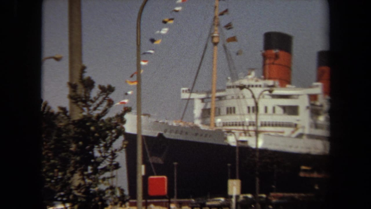 1971 8mm view of the Queen Mary docked in California, partly obscured by trees and dark foreground framing.
