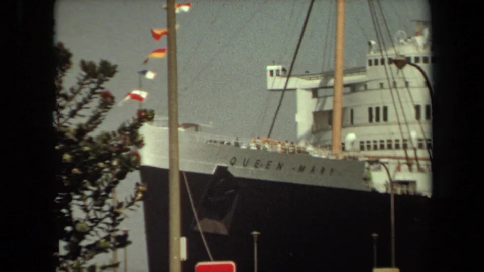 1971 8mm frame showing the Queen Mary bow and name lettering, partly blocked by a tree and vertical dockside pole.