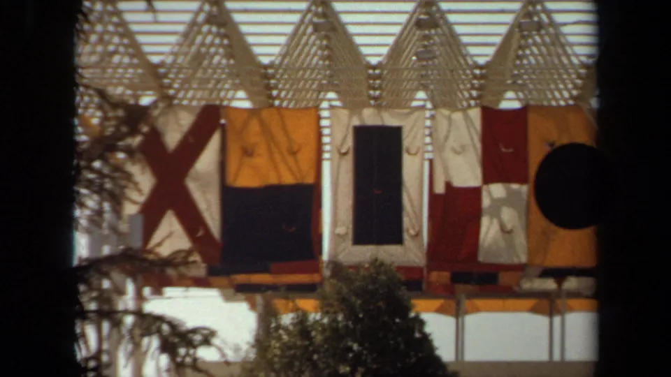 1971 8mm view of colorful nautical-style flags on a dockside structure near the Queen Mary setting in California.