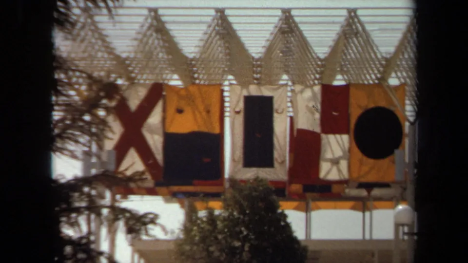 1971 Laguna Hills 8mm frame showing a close exterior view of a fort-like structure with large signal-style flags and trees in front.