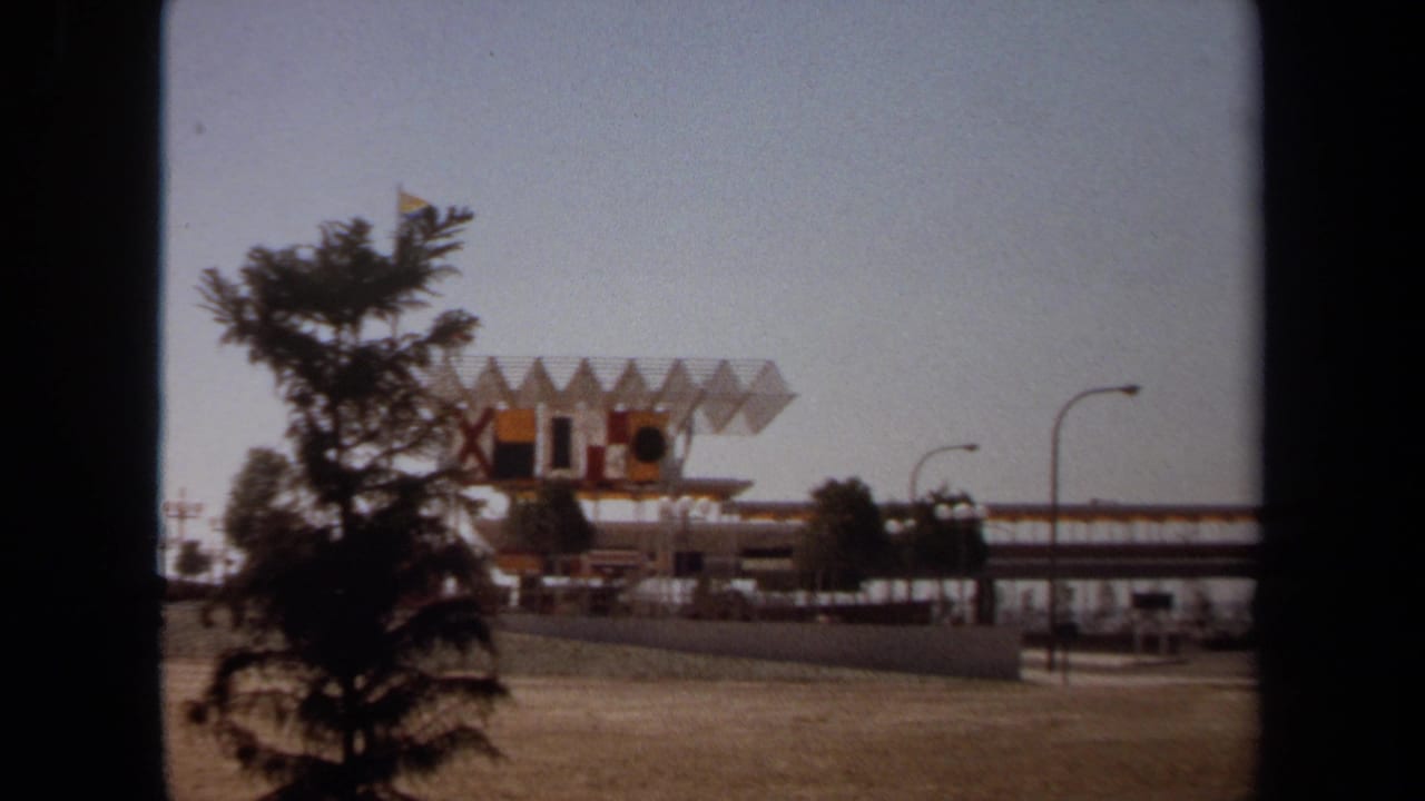 1971 8mm view of a California waterfront area with a geometric-roof building, road lamps, dry foreground grass, and dark framing at the edges.