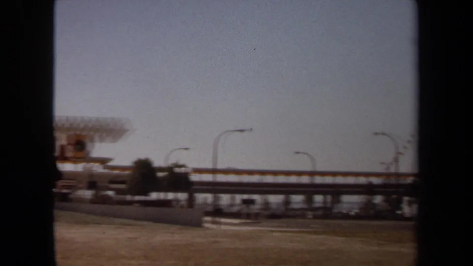 1971 8mm frame showing an elevated bridge or walkway across a waterfront setting, with curved lamps, low structures, and dark vertical framing.