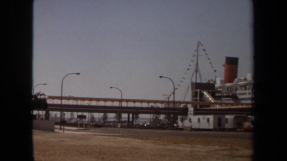 1971 8mm view of a large docked ocean liner beside an elevated walkway, with rigging, lamps, low buildings, and parked cars visible.
