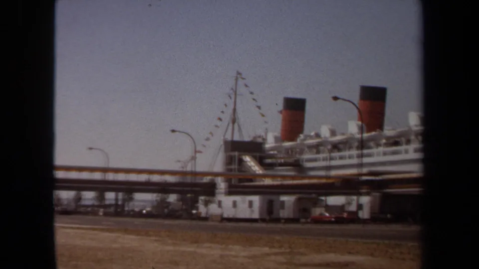 1971 8mm frame of a docked ocean liner with twin red-and-black funnels, rigging, lamps, parked cars, and dry foreground near the waterfront.