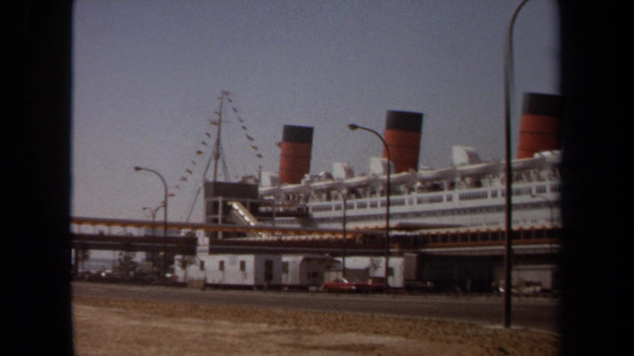 1971 8mm frame of a large white ocean liner at a port, seen from across a road with streetlights and dock structures in view.