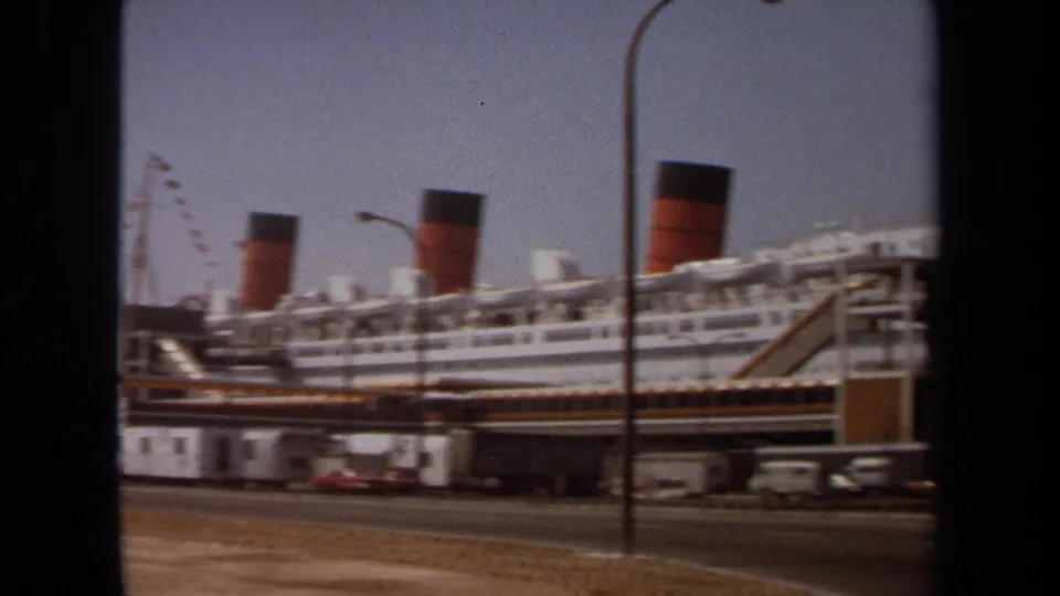 The ship remains docked in a 1971 home-movie view, framed by road traffic elements and a pier ramp in the foreground.