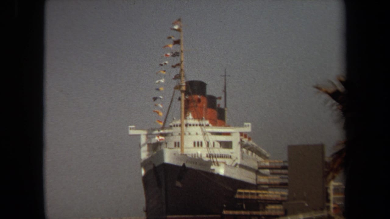 1971 Long Beach home-movie view of the Queen Mary bow, funnel, mast, and signal flags, partly framed by dark foreground edges.