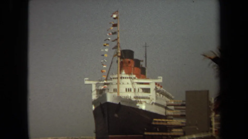 Queen Mary docked at Long Beach in 1971, with bow, funnel, mast, and flags visible through dark home-movie framing.