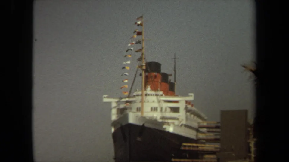 Soft 8mm view of the Queen Mary bow at Long Beach, with mast, signal flags, funnel, and dock elements visible.