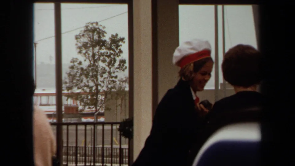 1971 Long Beach terminal moment showing a uniformed woman in a white hat speaking with a passenger or visitor near harbor-facing windows.