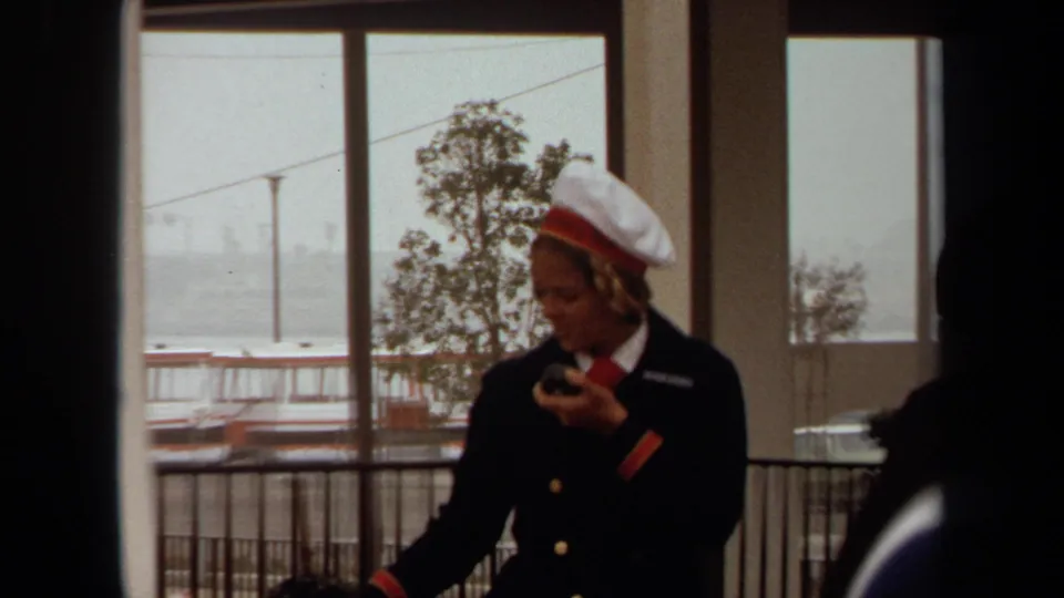 Uniformed woman at a Long Beach terminal looks down at a handheld microphone or radio, with windows and harbor railings behind her.