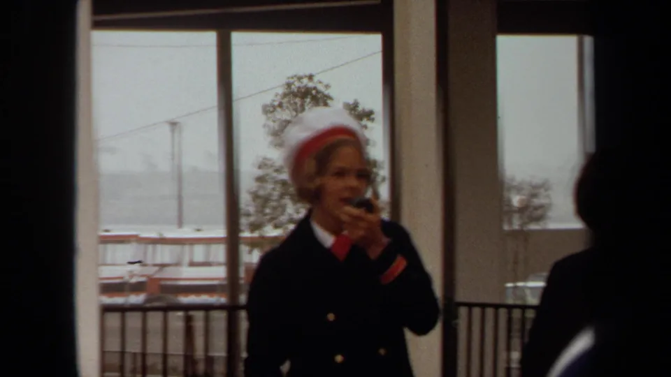 At a Long Beach cruise ship dock in 1971, a uniformed woman appears to make an announcement beside large windows overlooking the port.