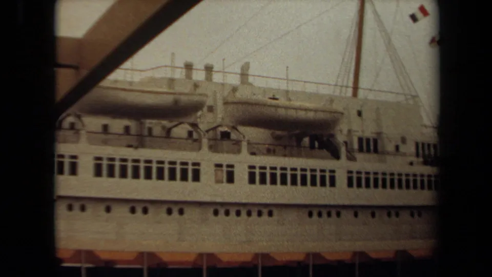 A white ocean liner at Long Beach is viewed from the dock area, showing lifeboats, deck railings, rows of windows, and mast rigging.
