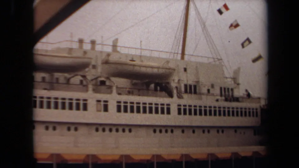 The camera holds on the docked passenger ship, with lifeboats and signal flags visible above rows of rectangular windows.