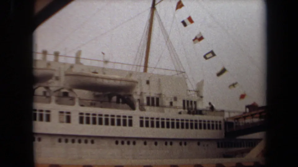 A dockside view of the Long Beach passenger ship shows its upper deck, lifeboats, mast rigging, and strings of flags in soft 8mm focus.