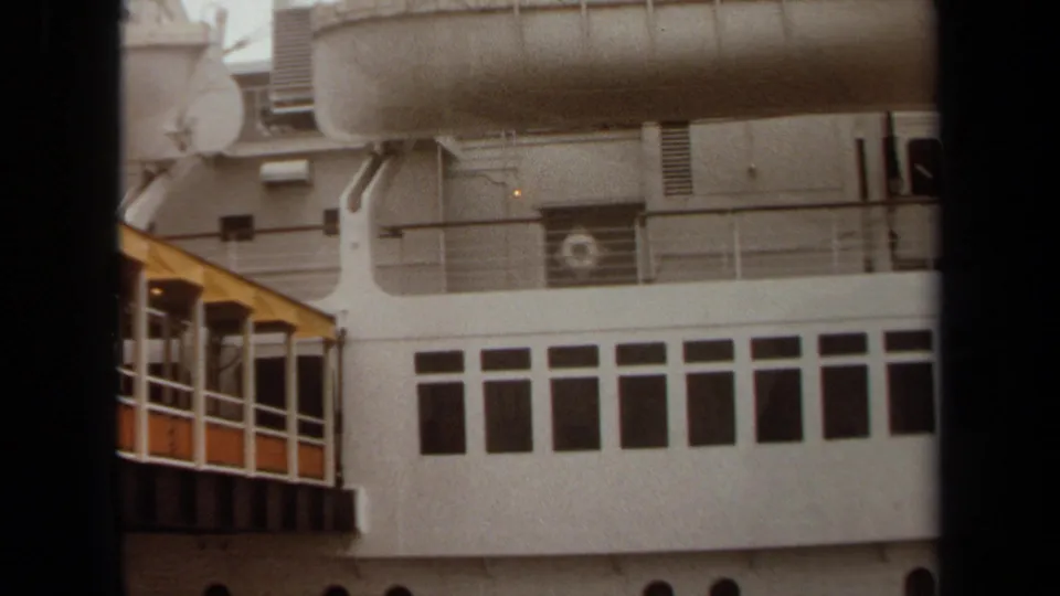1971 Long Beach frame of a docked ship showing a mounted lifeboat, upper railings, rectangular windows, and the edge of a yellow gangway.