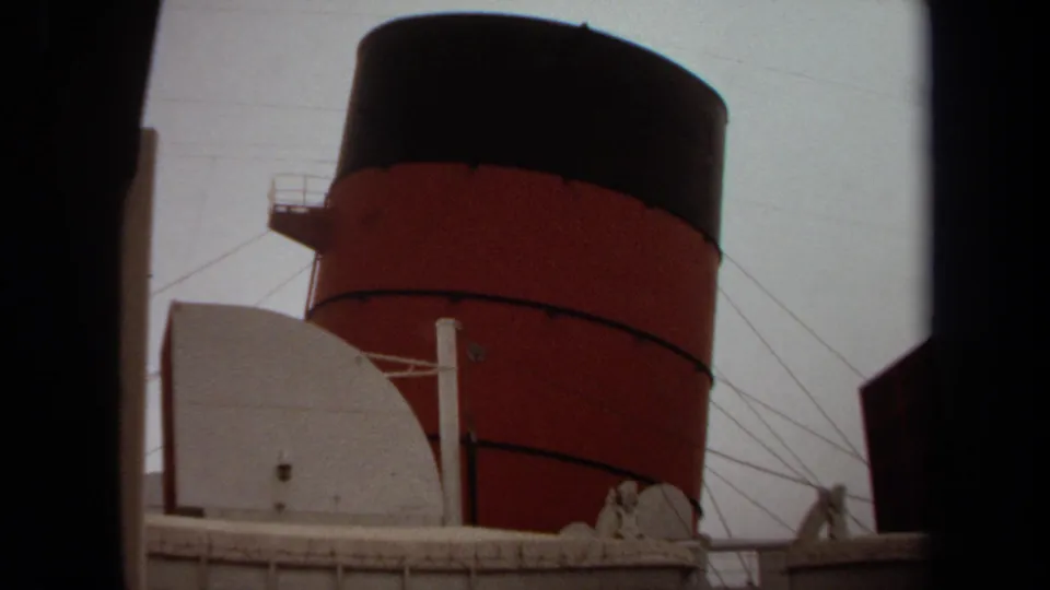 1971 Long Beach home-movie frame showing a red-and-black smokestack rising behind the ship's white structures and lifeboat.