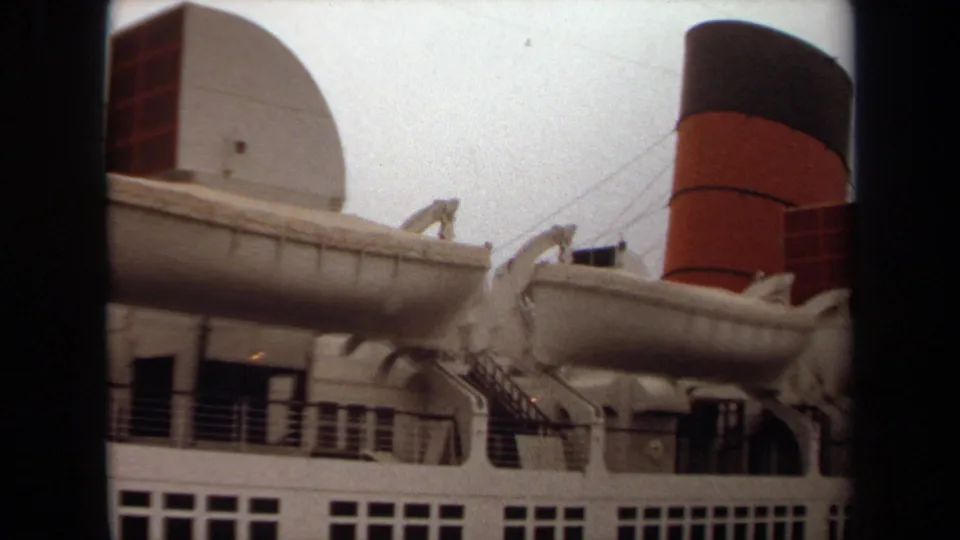 1971 Long Beach home-movie frame with the liner's port side, white lifeboat, rigging, railings, rows of windows, and red-and-black funnel.