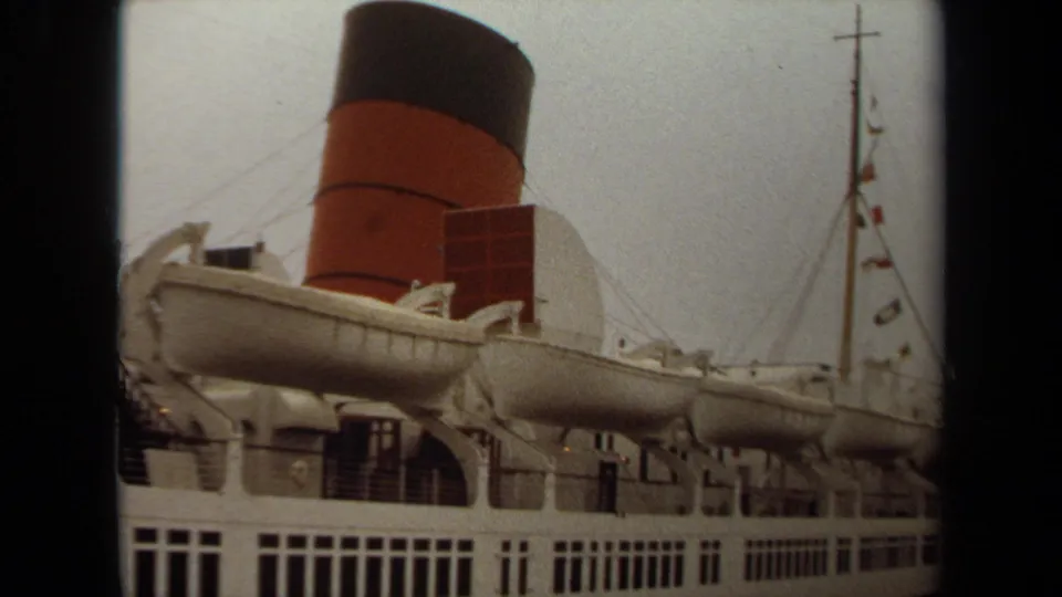 1971 Long Beach 8mm frame showing the ship's lifeboats, red-and-black funnel, rows of windows, and part of a mast with flags.
